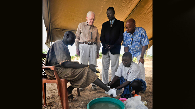 A volunteer treats a South Sudanese farmer with Guinea worm disease, flanked by former US President Jimmy Carter, Makoy Samuel Yibi, and Dr. Donald Hopkins, vice president of Health Programs at The Carter Center.